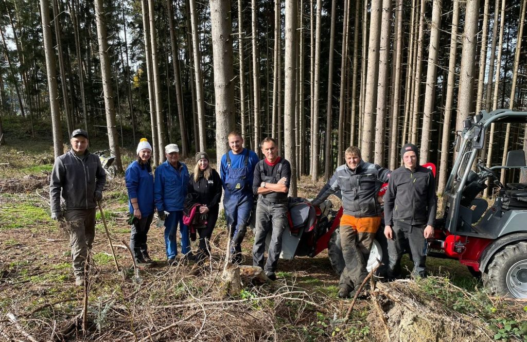 Eine Gruppe von Menschen, die neben einem Waldarbeitsfahrzeug im Wald stehen, lächeln und für ein Gruppenfoto posieren
