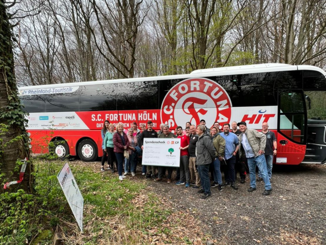 Eine Gruppe von Personen steht vor einem Reisebus mit dem Logo des S.C. Fortuna Köln, während sie gemeinsam ein Scheck-Symbol für eine Spendenaktion halten.