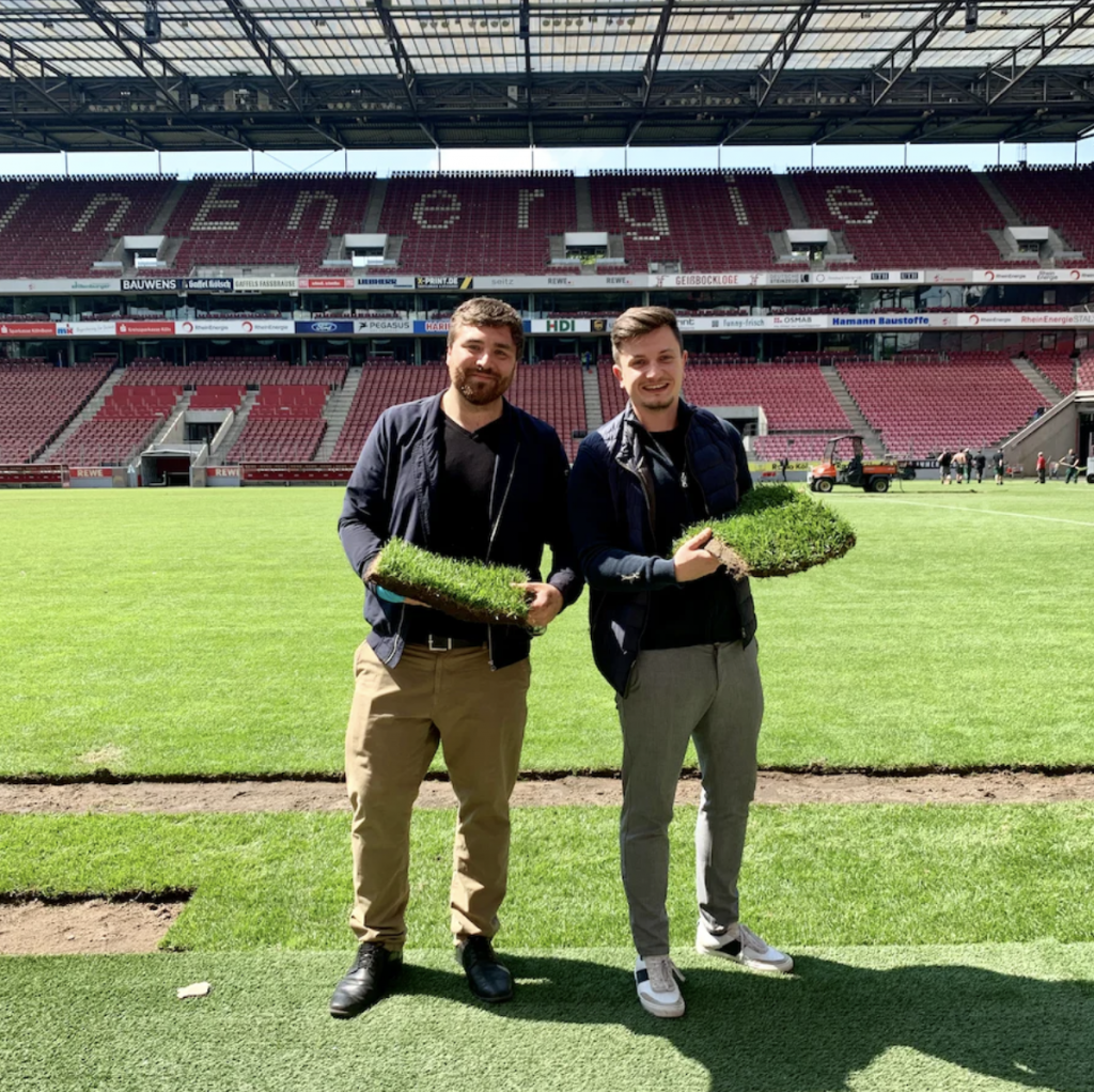 Die beiden Gründer Patrick Köhler und Robin Kracht stehen auf einem Fußballfeld und halten Stücke von Rasen in den Händen, im Hintergrund sind die Tribünen eines Stadions zu sehen