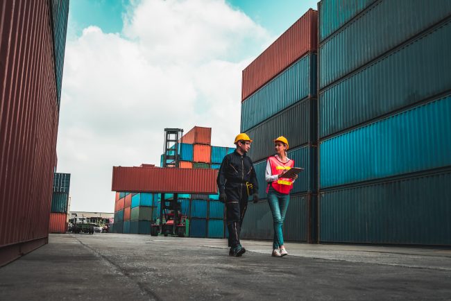 Zwei Personen mit Schutzhelmen, die in einem Containerhafen gehen und Unterlagen besprechen, symbolisieren das Management des CO2-Fußabdrucks in der Logistikbranche.