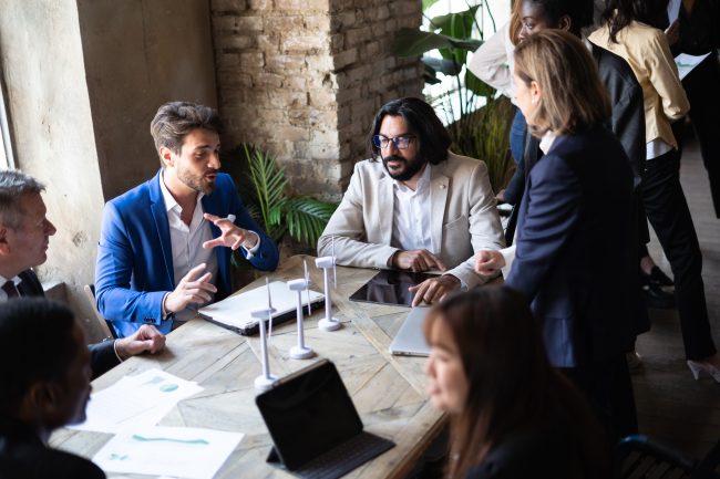 CO2 Reduktion Unternehmen - Eine Gruppe von professionell gekleideten Menschen führt eine lebhafte Diskussion an einem Konferenztisch in einem Büro mit Ziegelwandhintergrund